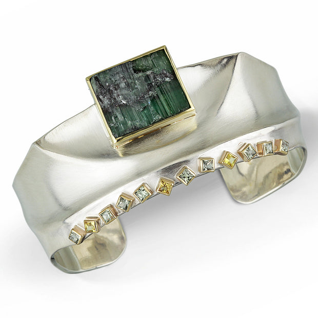 Full view of Green Tourmaline and Fancy Colored Sapphire - Ridge Cuff. This is a silver cuff that showcases a square piece of raw green tourmaline encompassed in gold that hangs a little off the cuff. On the opposing side are twelve fancy colored princess cut sapphires.