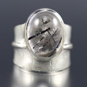 Full view of Tourmalated Quartz Ridge Ring. This ring showcases an ovular cut piece of tormalated quartz on a thick silver band that resembles the spine of a leaf.