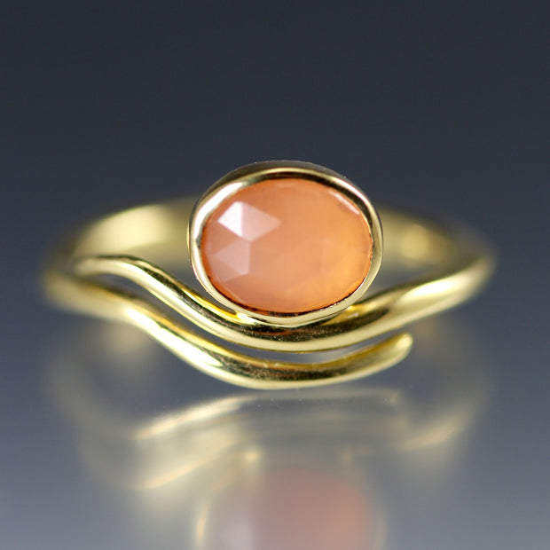 Full view of Peach Moonstone Wave Ring. The peach moonstone is cut in an oval and lays on a gold band that does not fully connect but creates the illusion of a wave where the moonstone is with two lines of gold wire bending in the shape of a swivel.