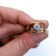 Full view of Andromeda Ring being held in hand to help give an idea of its scale. This ring is made of yellow gold with a set cornflower sapphire at its top and accents of silver and diamonds throughout its open natured band.