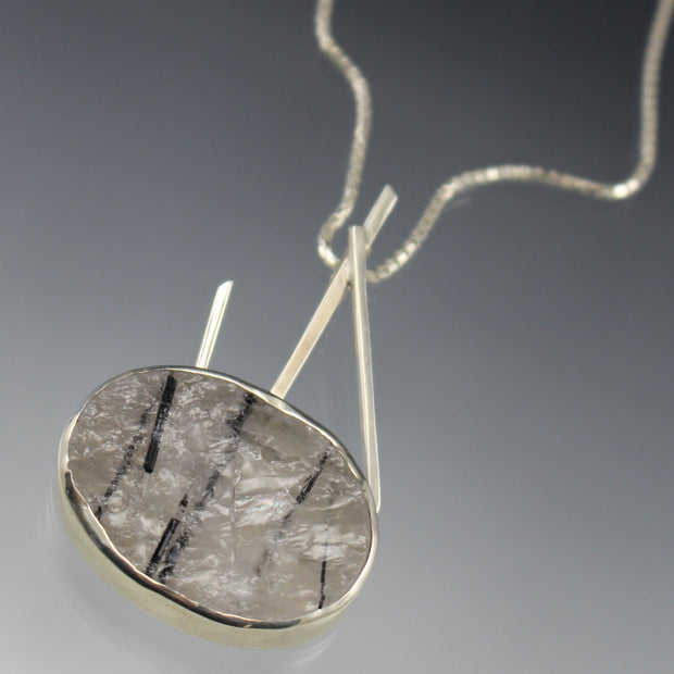 Full image of pendant on Oval Tourmalated Quartz - Sticks and Stones Pendant. This pendant showcases an ovular cut raw tourmalated quarts set in silver. There are three pieces of silver square wire that protrude out of the top of the quartz. The left-most wire is angled towards the left and the two on the ring make an "A" shape and hook for the chain to run through.