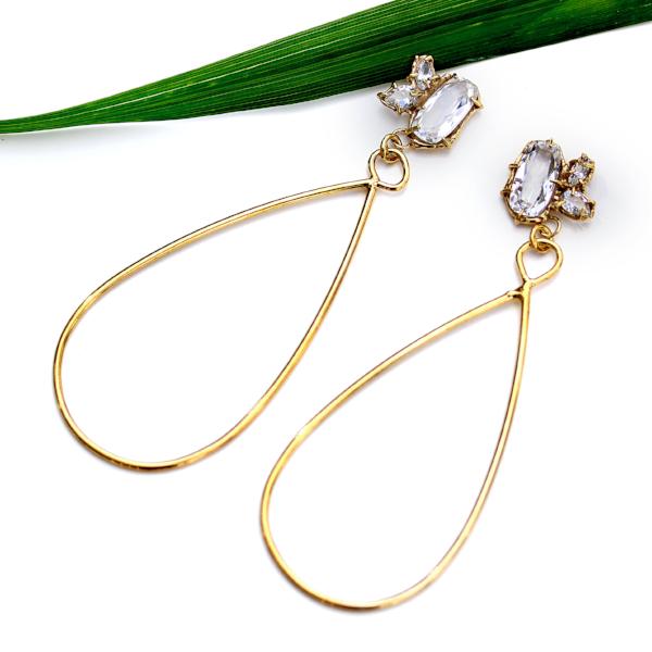Gold dangle earrings with three white topaz gemstone in various shapes and a tear drop shaped dangle with a green plant leaf in the background.