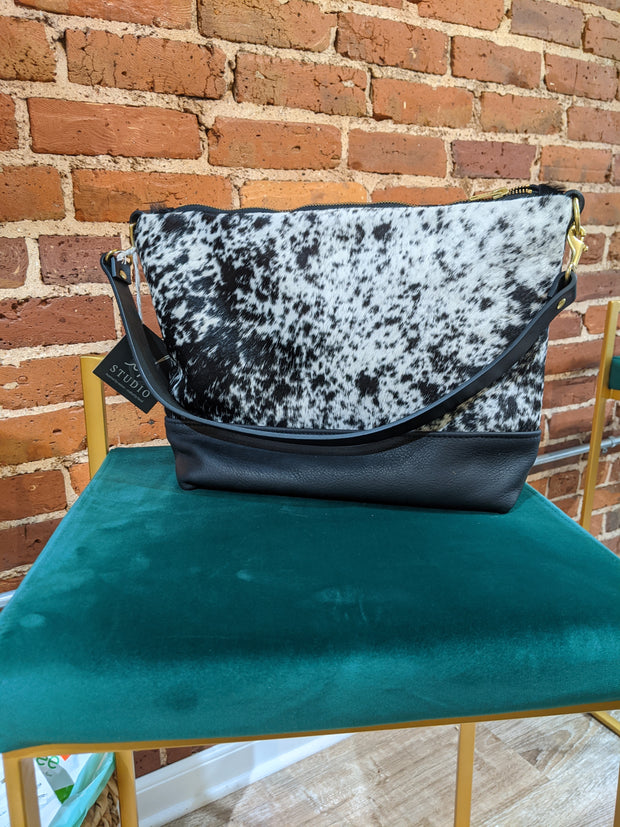 Full image of Virginia shoulder bag in a black and white cowhide print sitting on chair. Has black leather accents on bottom of bag and straps.