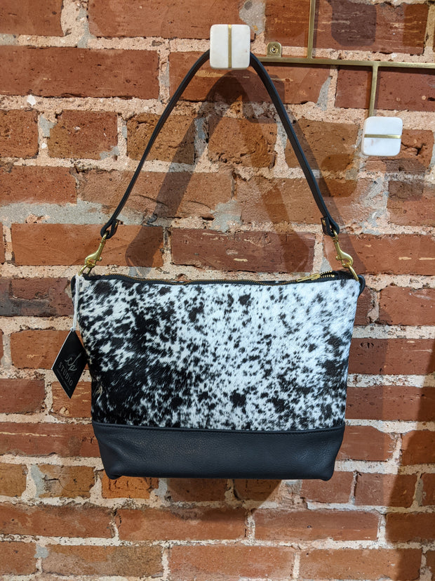 Full image of Virginia shoulder bag in a black and white cowhide print. hanging from hook Has black leather accents on bottom of bag and straps.