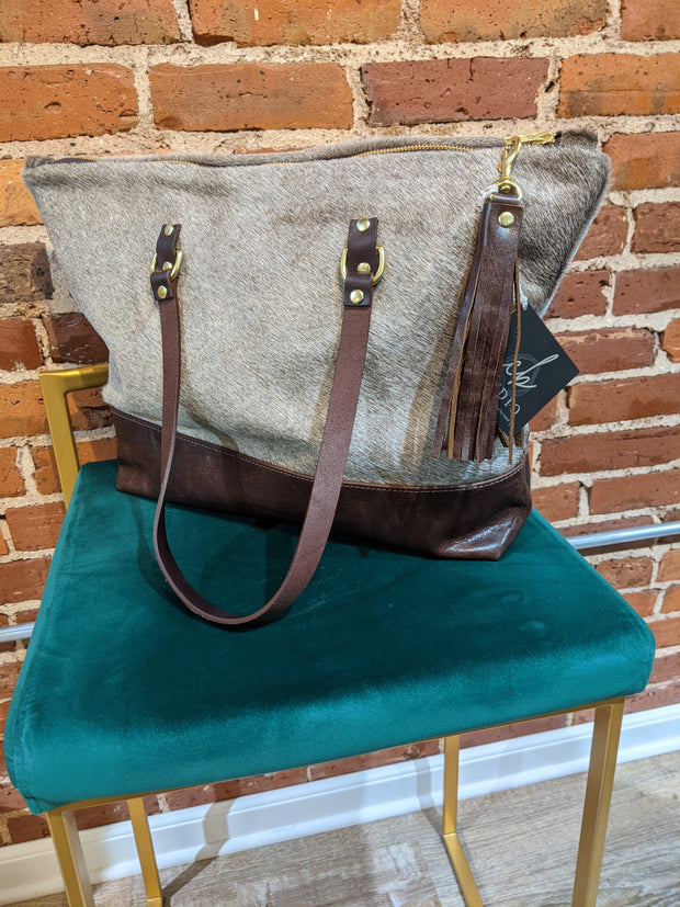Full image of Ashlyn Tote Bag. This bag looks like it is made from cowhide and has leather looking accents for the bottom of the tote and the two handles.