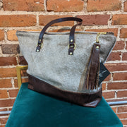 Full image of Ashlyn Tote Bag. This bag looks like it is made from cowhide and has leather looking accents for the bottom of the tote and the two handles.