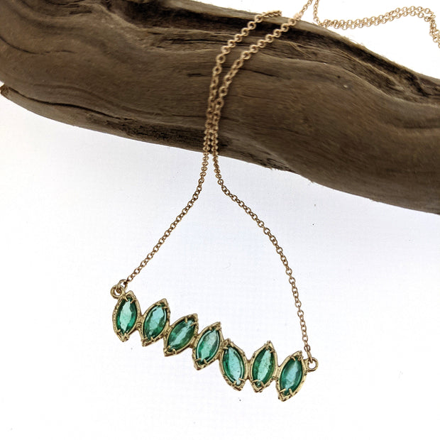 Full view of Emerald Cherin Necklace hanging off of branch. Seven Sparkling natural marquise shaped Emeralds delicately adorn this irregular line-style bar necklace with subtle organic detailing. 