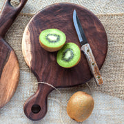 Full view of 8" Round Walnut Cutting Board being used to cut a kiwi. In the shape of a circle with a handle.