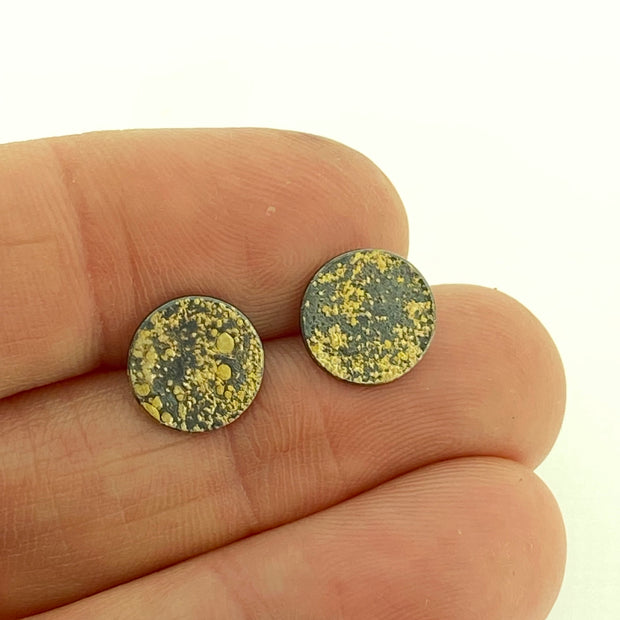 Full view of Lichen Moon Earring in-between two finger to help give an idea of its scale. These studs are in the shape of circle, made of oxidized silver and have gold accents.