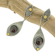 Full view of Fused Botanical Dot - Garnet Earrings hanging off of a stick. These earrings are in the shape of a leaf at the top (made of textured oxidized silver) with a gold dot and a hinge going through. Attached to the hinge is a bigger oxidized leaf with a set garnet and one dot of gold at the tip.