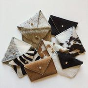 Full image of nine different card wallets that all have a different fur (cowhide, zebra, etc.). Each one has an accent of a brass bead that holds the lid of the card holder closed.