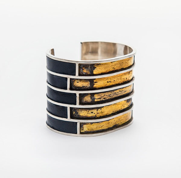 Full view of A.G. Cuff. This large cuff is split up into five horizontal rectangles with silver wire. The sides of the cuff are inlaid with black leather and the front five rectangle are brushed with gold on top of an oxidized surface.