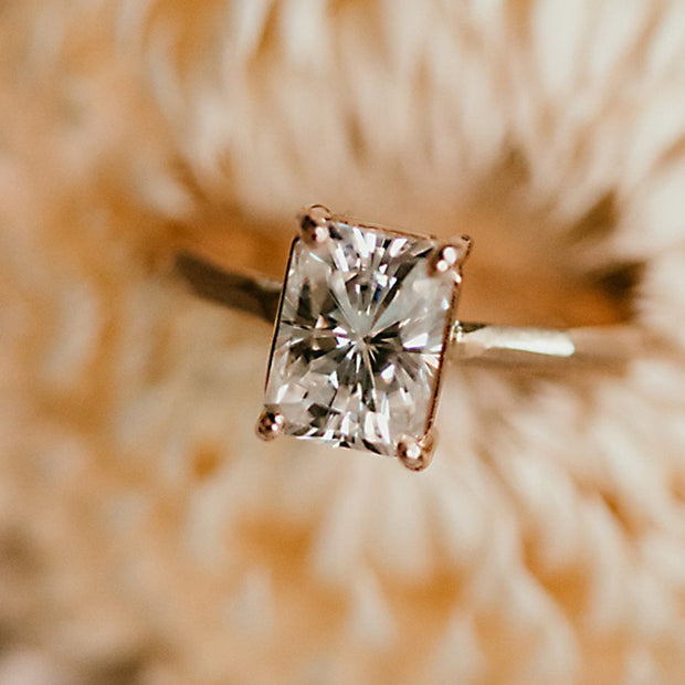 Detail image of set moissanite on Radiant Cut Moissanite - Chiseled Engagement Ring. This ring lays on a gold band that has broad facets that resemble rock formations. A satin finish on the band and a high polish on the stone setting sets off this gorgeous stone. The moissanite is cut in the shape of a rectangle.