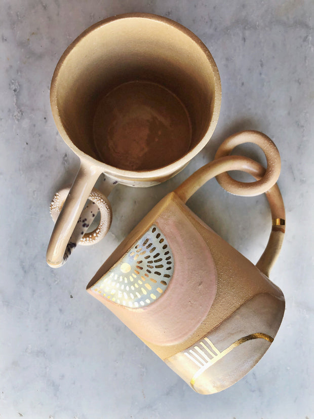 Full image of two Ring mug made with a variety of creams, tans, and whites. One mug is laying down and the other we can see inside of the mug. There is a circle ring that dangles from the handle of the mug.