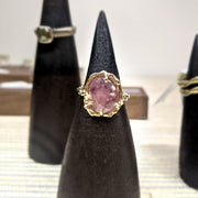 Full view of Roxanna Ring - Large on black pedestal. The Roxanna features organic carved metalwork with a delicately dappled surface.     A large 1.4 asymmetrical shaped Umba Sapphire in a beautiful shade of pale pink is set in 14K (recycled) Yellow Gold. 