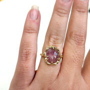 Full view of Roxanna Ring - Large on a woman's hand to help give an idea of its scale. The Roxanna features organic carved metalwork with a delicately dappled surface.     A large 1.4 asymmetrical shaped Umba Sapphire in a beautiful shade of pale pink is set in 14K (recycled) Yellow Gold. 