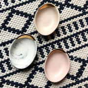 Full view of three small oval dishes. Two are in matte pink with the top fifth dipped in gold and the third in a black and white marble with the top fifth dipped in gold.